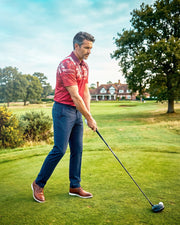 A man in a red polo shirt and navy trousers stands on a golf course, holding a golf club and preparing to swing. Trees, grass, and a clubhouse are visible in the background.