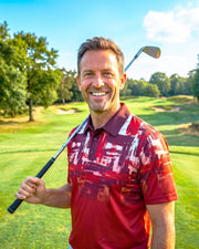 A smiling man on a golf course holding a golf club, wearing a red and white patterned short-sleeve polo shirt.