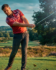 A male golfer in a red patterned polo shirt and dark trousers swings a golf club on a sunny golf course, with trees and grass in the background.