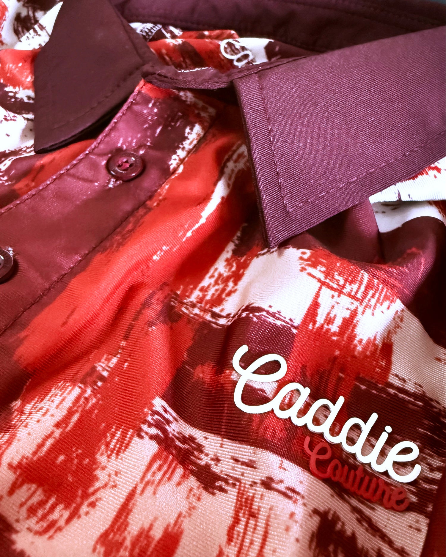 Close-up of a collared button-up shirt with a red, white, and dark burgundy abstract print. The shirt features a solid burgundy collar and button placket, with the brand name 'Caddie Couture' embroidered in white script on the chest.