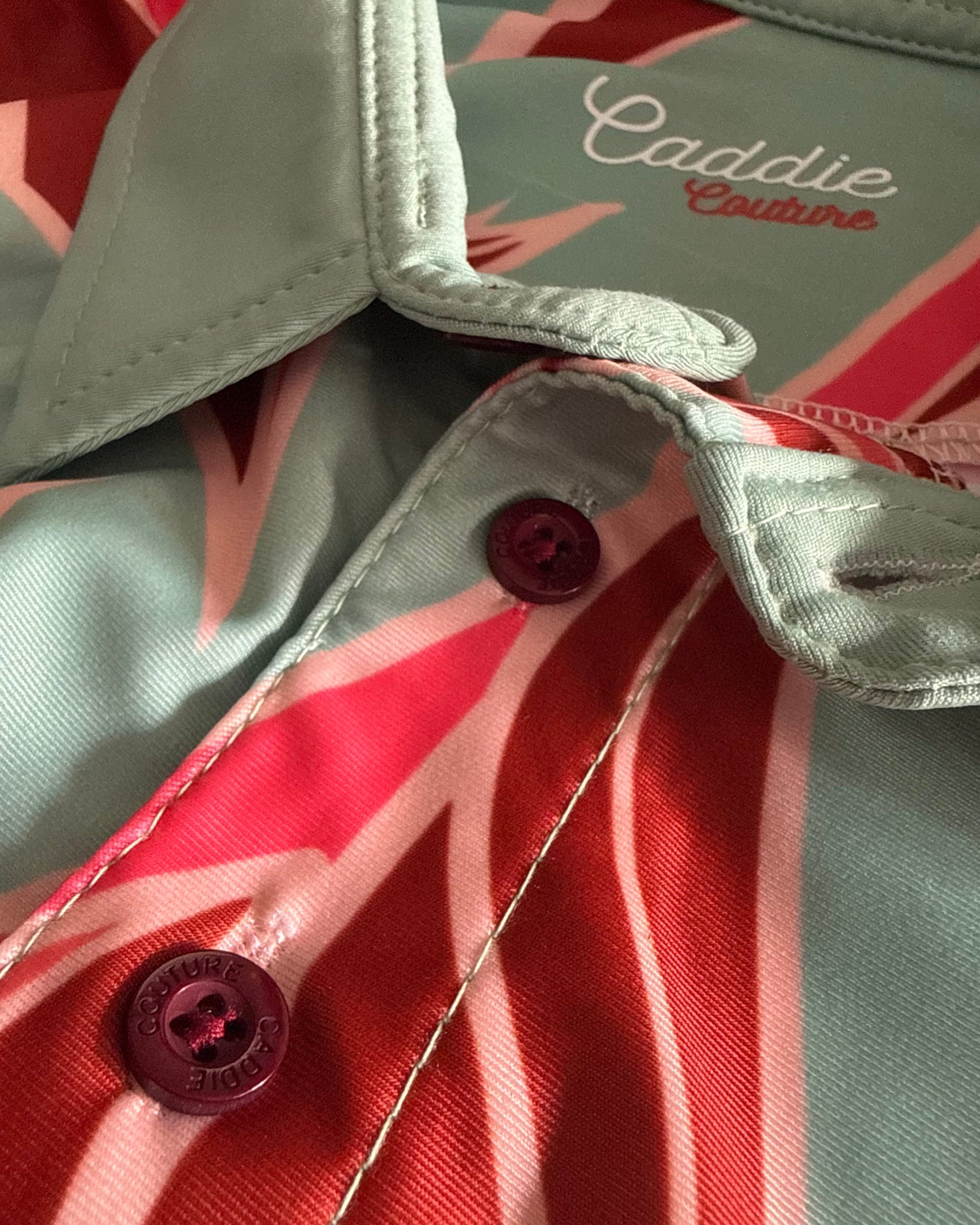 Close-up of a Caddie Couture button-down shirt featuring a tropical palm leaf pattern in sage green, coral red, and pink, with dark red buttons.