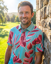 A smiling man wearing a turquoise short-sleeve polo shirt with a bold pink and red leaf pattern, leaning against a rustic stone wall on a sunny golf course.