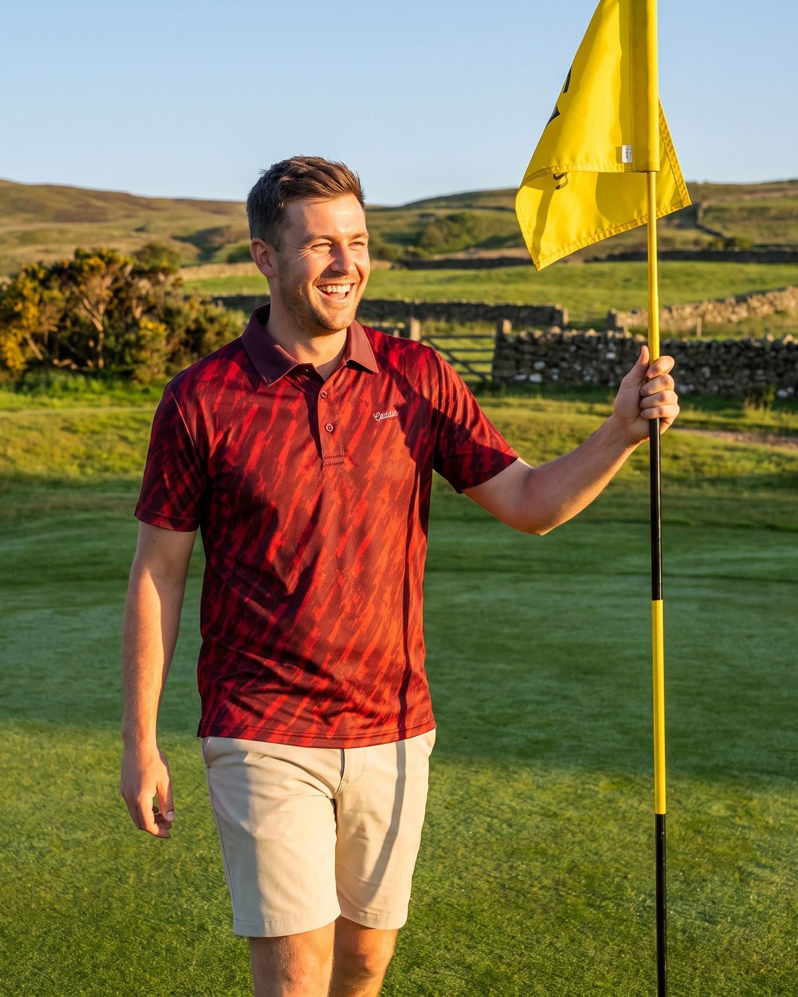 A smiling man on a golf course wearing a red patterned polo shirt and beige shorts, holding a yellow flag. The background shows green grass, stone walls, and hills under a clear blue sky.