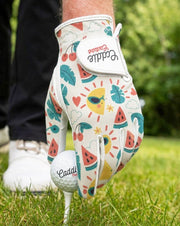 Colorful golf glove with fruit patterns and a matching golf ball on a tee, set against a blurred green outdoor background.