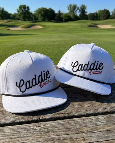 Two white caps with 'Caddie Couture' text on a wooden surface with a golf course background.