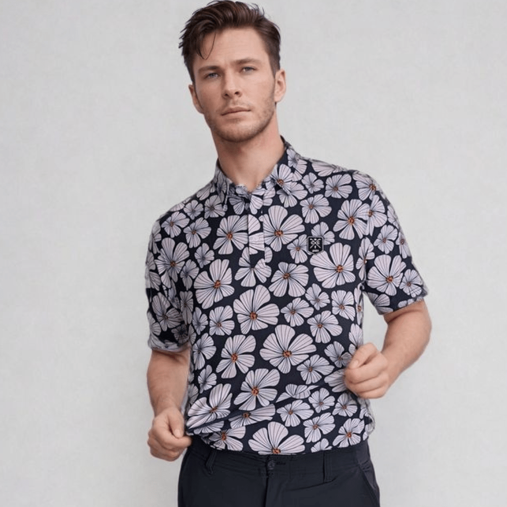 A man wearing a black golf polo with a floral pattern consisting of pink and white flowers and green leaves.